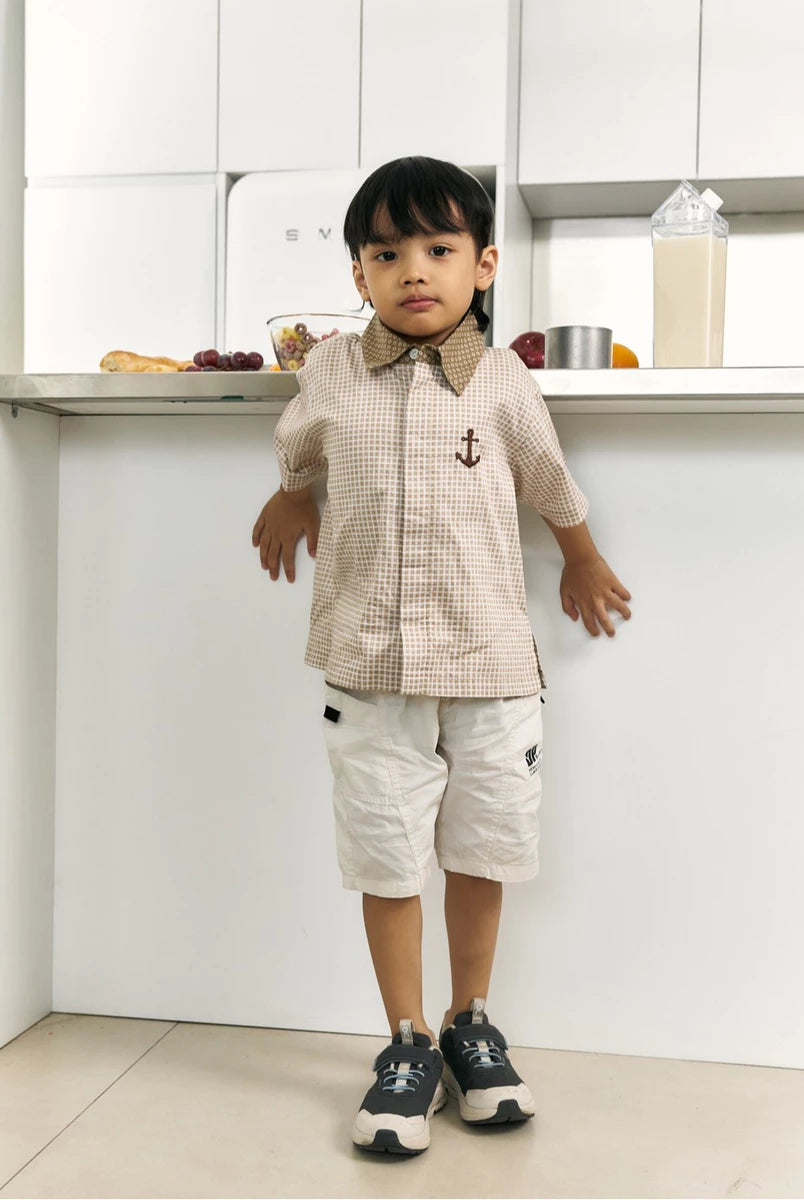 A young boy stands in a bright, modern kitchen wearing a beige and white checkered short-sleeve button-up shirt by Studio Tui with a brown collar and a small anchor emblem, paired with off-white shorts and dark sneakers.