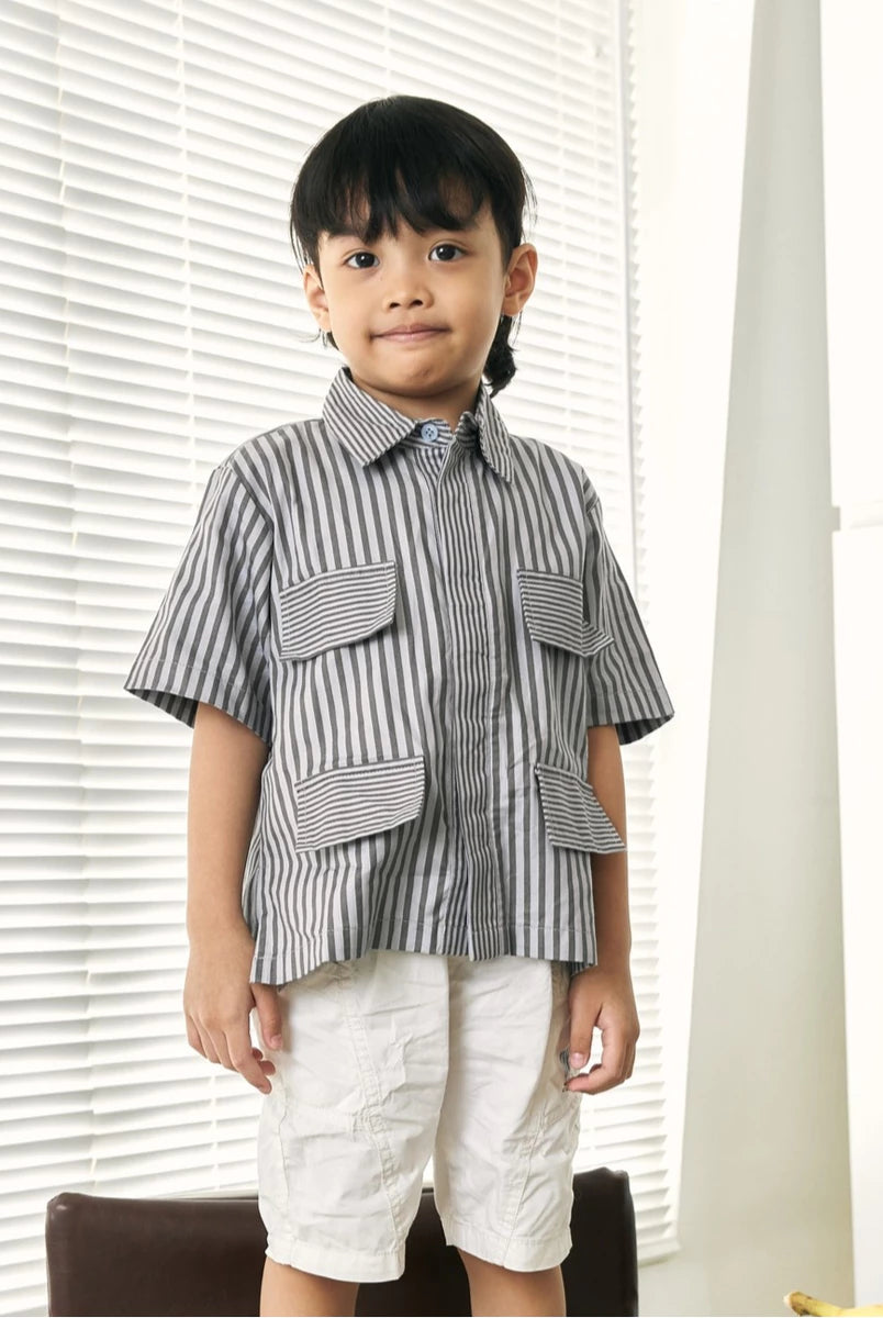 A young boy wearing a short-sleeve button-up shirt by Studio Tui with vertical gray and white stripes, featuring large front flap pockets, paired with off-white shorts, standing in front of white window blinds.