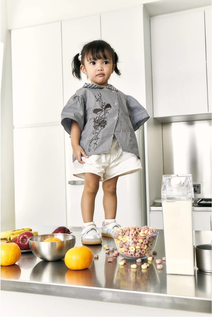 A toddler girl stands in a modern white kitchen wearing a gray and white striped smock top by Studio Tui with a dark floral print, white shorts, and white sneakers. In the foreground on a stainless steel counter are bowls of fruit, cereal, and a carton.