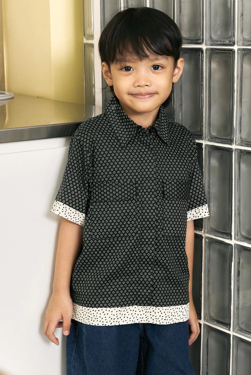 A young boy wearing a short-sleeve black button-up shirt by Studio Tui with a white geometric pattern and contrasting white cuffs and hem trim, paired with dark blue jeans, standing in front of a glass block wall.