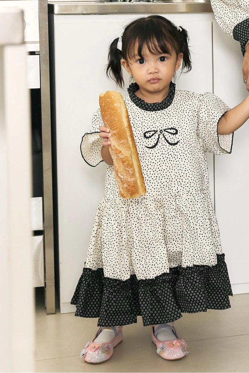 A young girl with dark hair in pigtails stands indoors holding a baguette. She is wearing a cream-colored dress by Studio Tuiwith black polka dots, black ruffled trim at the collar and hem, and a black bow motif on the chest.