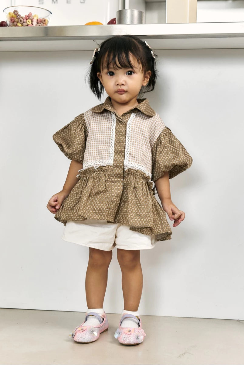A toddler girl wearing a brown and beige patterned short-sleeve top by Studio Tui with white shorts and pink shoes, standing in front of a white wall.
