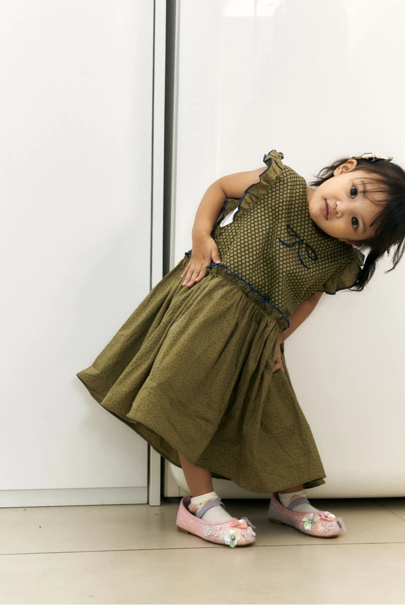 A toddler girl wearing an olive green sleeveless dress by Studio Tui with a polka dot bodice, ruffled shoulders, and a flared skirt, posing against a white wall.