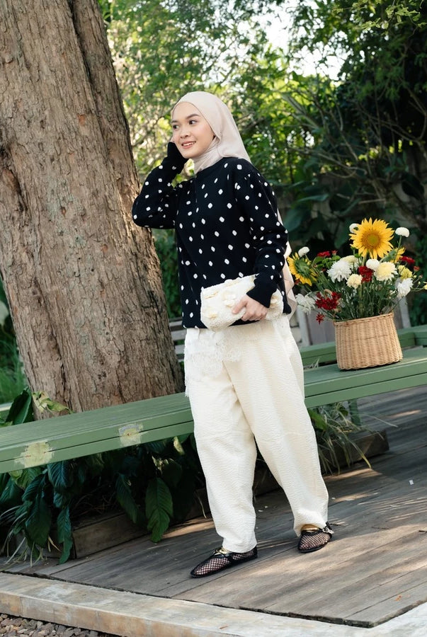 A woman wearing a black long-sleeve sweater by Haidee Orlin with white polka dots and cream-colored wide-leg trousers, standing on a wooden deck next to a large tree trunk with a green bench and a basket of flowers in the background.