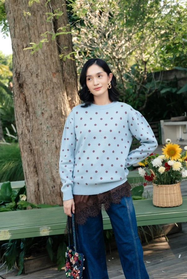 A woman stands outdoors in a garden setting wearing a light blue crewneck sweatshirt by Haidee Orlin with a small brown polka dot pattern, paired with dark blue wide-leg jeans and a brown lace hem detail. She is holding a small floral handbag next to a green bench with a basket of flowers.