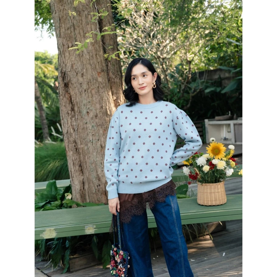 A woman stands outdoors in a garden setting wearing a light blue crewneck sweatshirt by Haidee Orlin with a small brown polka dot pattern, paired with dark blue wide-leg jeans and a brown lace hem detail. She is holding a small floral handbag next to a green bench with a basket of flowers.
