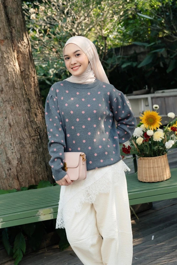 A woman wearing a dusty blue-gray long-sleeve knit sweater by Haidee Orlin with pink polka dots, a light beige hijab, and white lace-trimmed pants, standing outdoors next to a green bench with a flower basket.