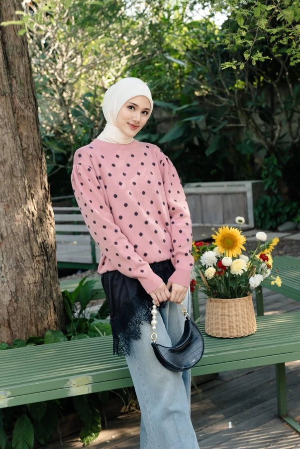 A woman wearing a pink long-sleeve sweater by Haidee Orlin with black polka dots, a white hijab, and light blue jeans stands outdoors on a green bench holding a black crescent handbag. A basket of flowers sits on the bench beside her, with trees and garden foliage in the background.