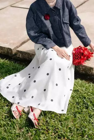 Person wearing a denim jacket and white polka dot skirt by Haidee Orlin holding red flowers on grass.