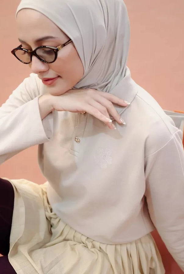 A woman wearing a light beige hijab and a matching long-sleeve polo shirt by Haidee Orlin with a pleated skirt, posed against a soft peach background.