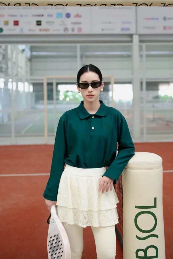 A woman stands on an indoor tennis court wearing an emerald green long-sleeve polo shirt by Haidee Orlin, a white tiered skirt, white tights, and black sunglasses, holding a tennis racket next to a BNSOL bollard.