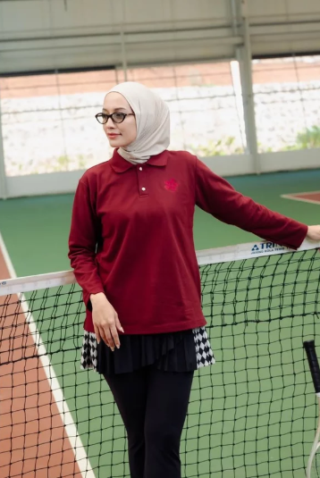 A woman wearing a hijab and a long-sleeve polo shirt in deep red by Haidee Orlin, standing on an indoor tennis court behind a net.