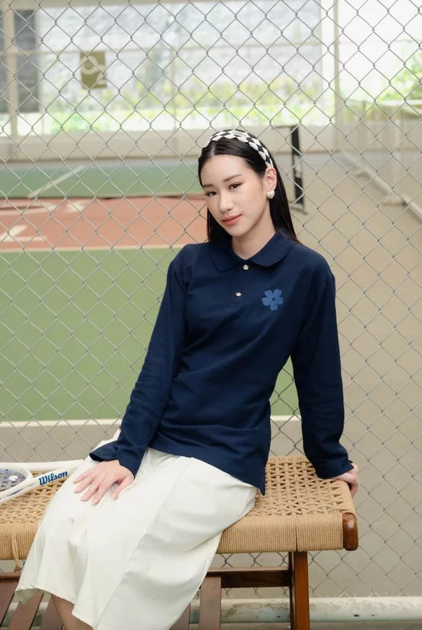 A woman sits on a woven bench in front of a chain-link fence at an outdoor sports field. She is wearing a navy blue long-sleeve polo shirt by Haidee Orlin with a small blue floral embroidery on the chest, paired with a cream-colored skirt.