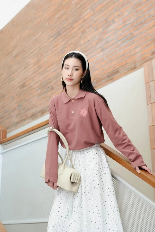 A woman stands on an indoor staircase wearing a dusty rose long-sleeve polo shirt by Haidee Orlin with a small pink flower detail on the chest, paired with a white polka-dot skirt and carrying a cream-colored shoulder bag.