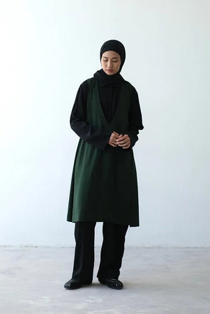 A person wearing a long green vest by Rashawl with a round neckline, sleeves, and a split at the back, paired with dark pants and black shoes.