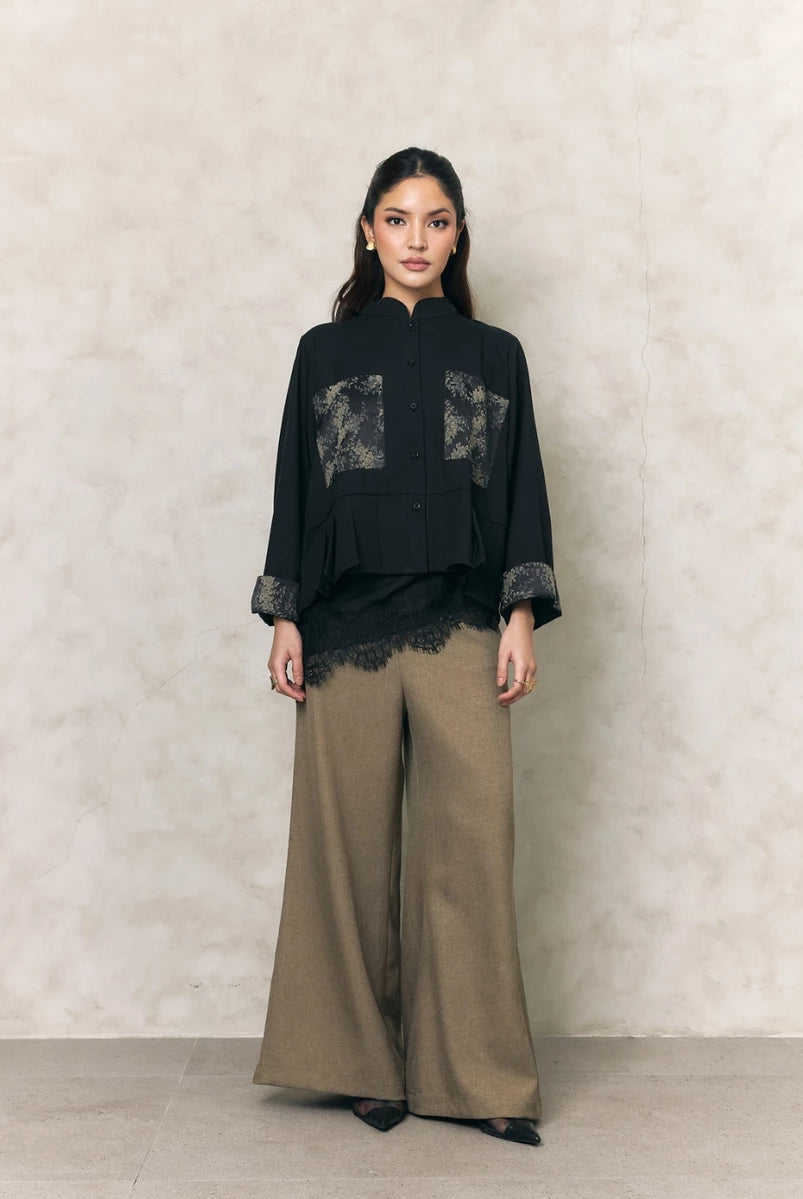 Woman wearing a black shirt with lace details by Studio Tui and beige pants against a neutral background