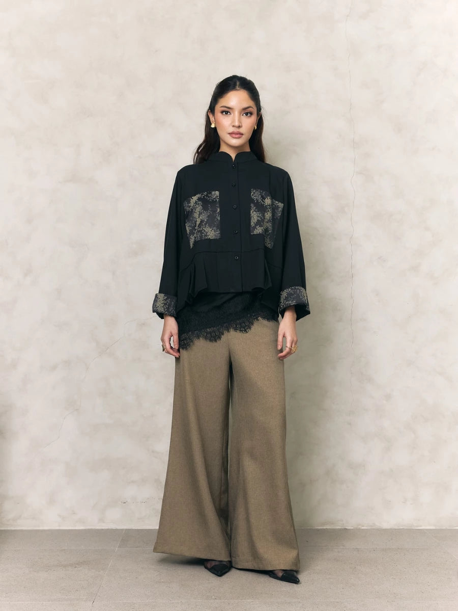 Woman wearing a black shirt with lace details by Studio Tui and beige pants against a neutral background