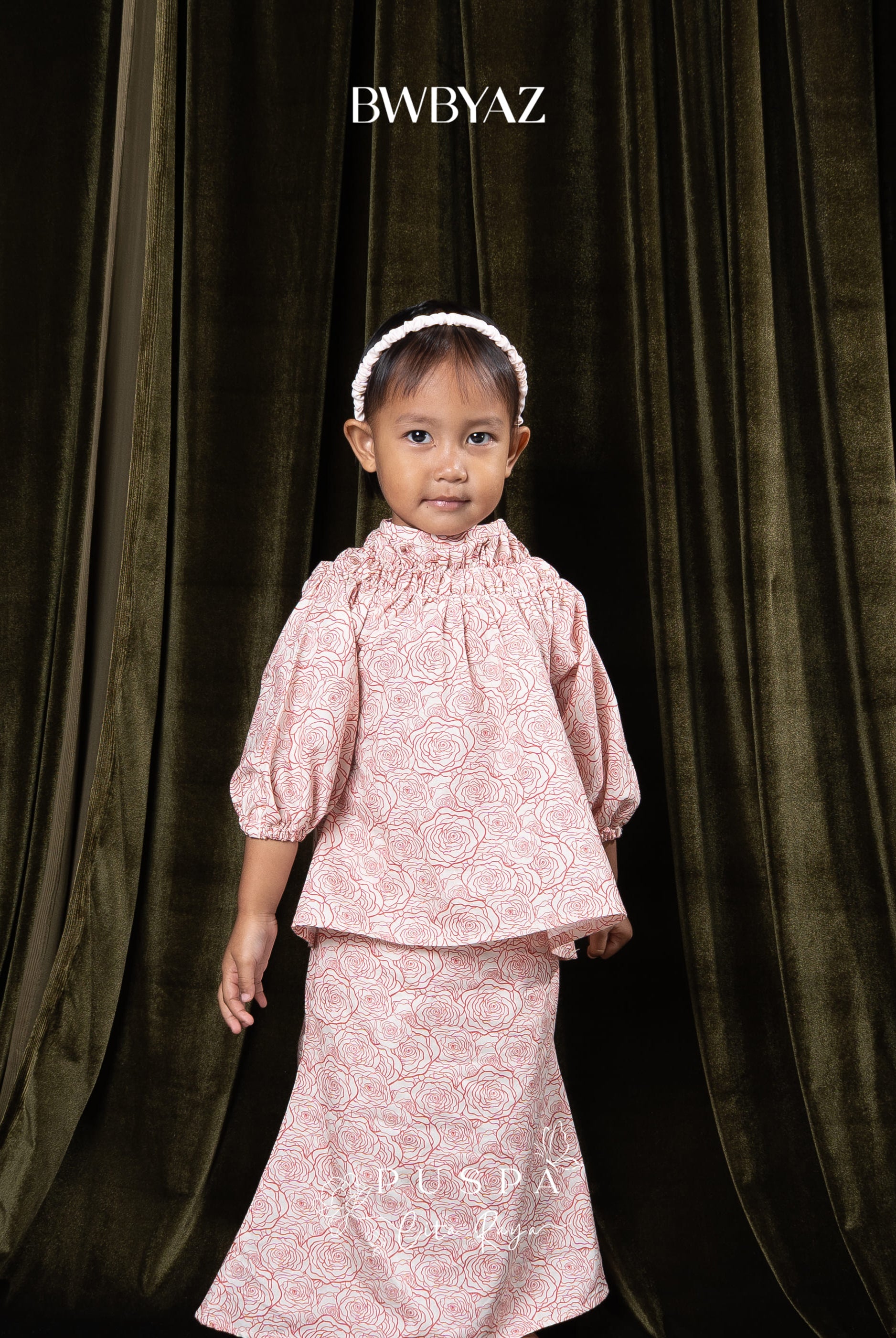 Child wearing a pink patterned top and skirt with a dark curtain background and 'BWBYAZ' branding.