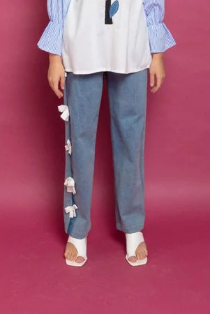 A person standing against a purple background, wearing blue denim pants with decorative white flowers on the leg, paired with a white shirt and beige shoes.
Seorang wanita berdiri dengan latar belakang ungu, mengenakan celana denim biru dengan hiasan bunga-bunga putih di bagian kaki, dipadukan dengan kemeja putih dan sepatu krem.