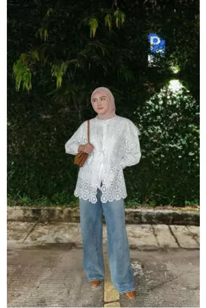 A person wearing a white vest by Abame with a floral pattern, blue jeans, and brown shoes, standing on a sidewalk with greenery in the background.