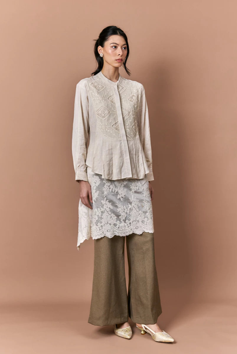 Woman wearing a beige embroidered shirt by Studio Tui, lace skirt, and brown pants against a beige background