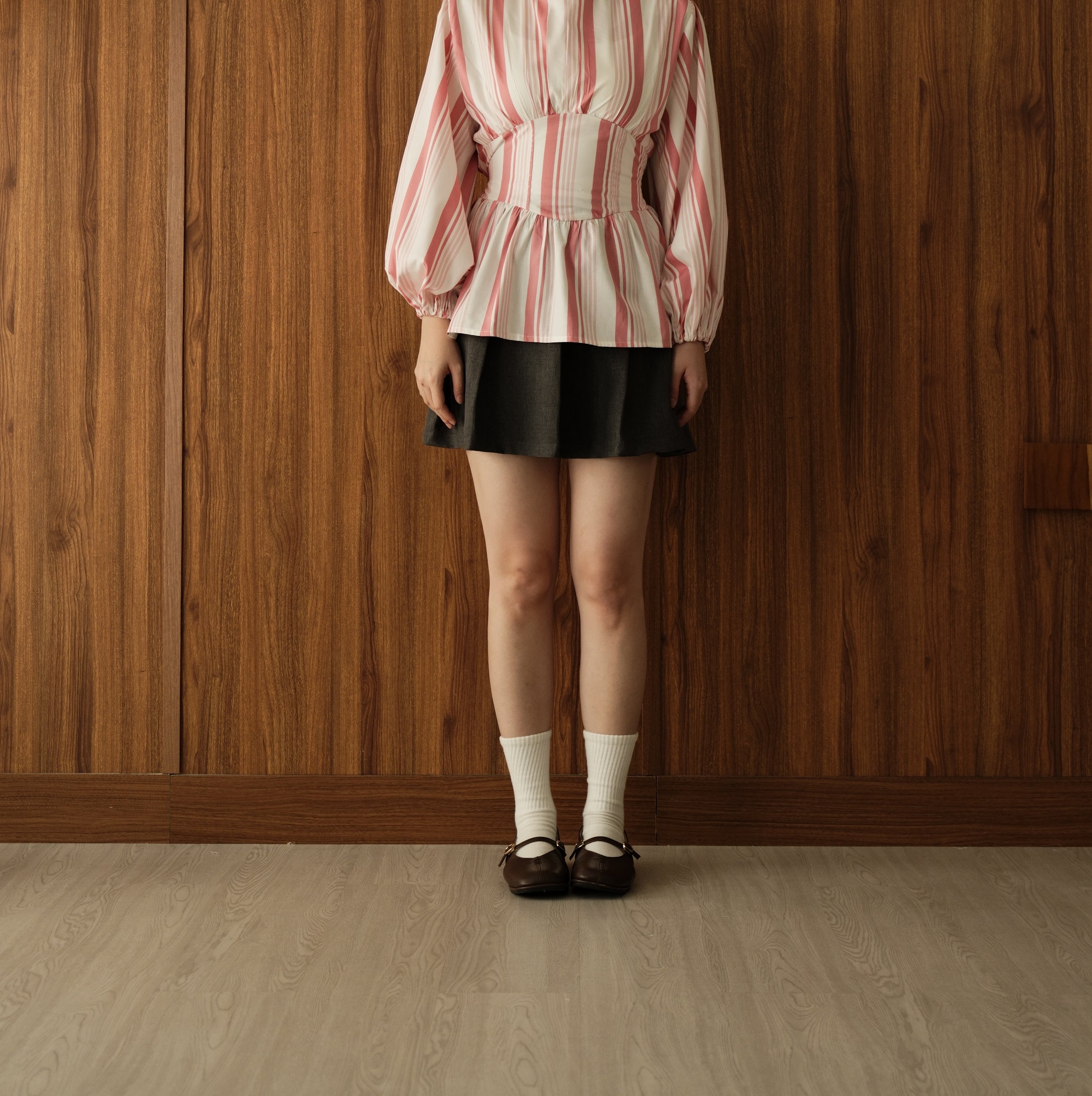 A young woman stands against a wood-paneled wall wearing a pink and white vertically striped long-sleeve blouse by BWBYAZ with a peplum waist, a black pleated mini skirt, white socks, and dark brown Mary Jane shoes.