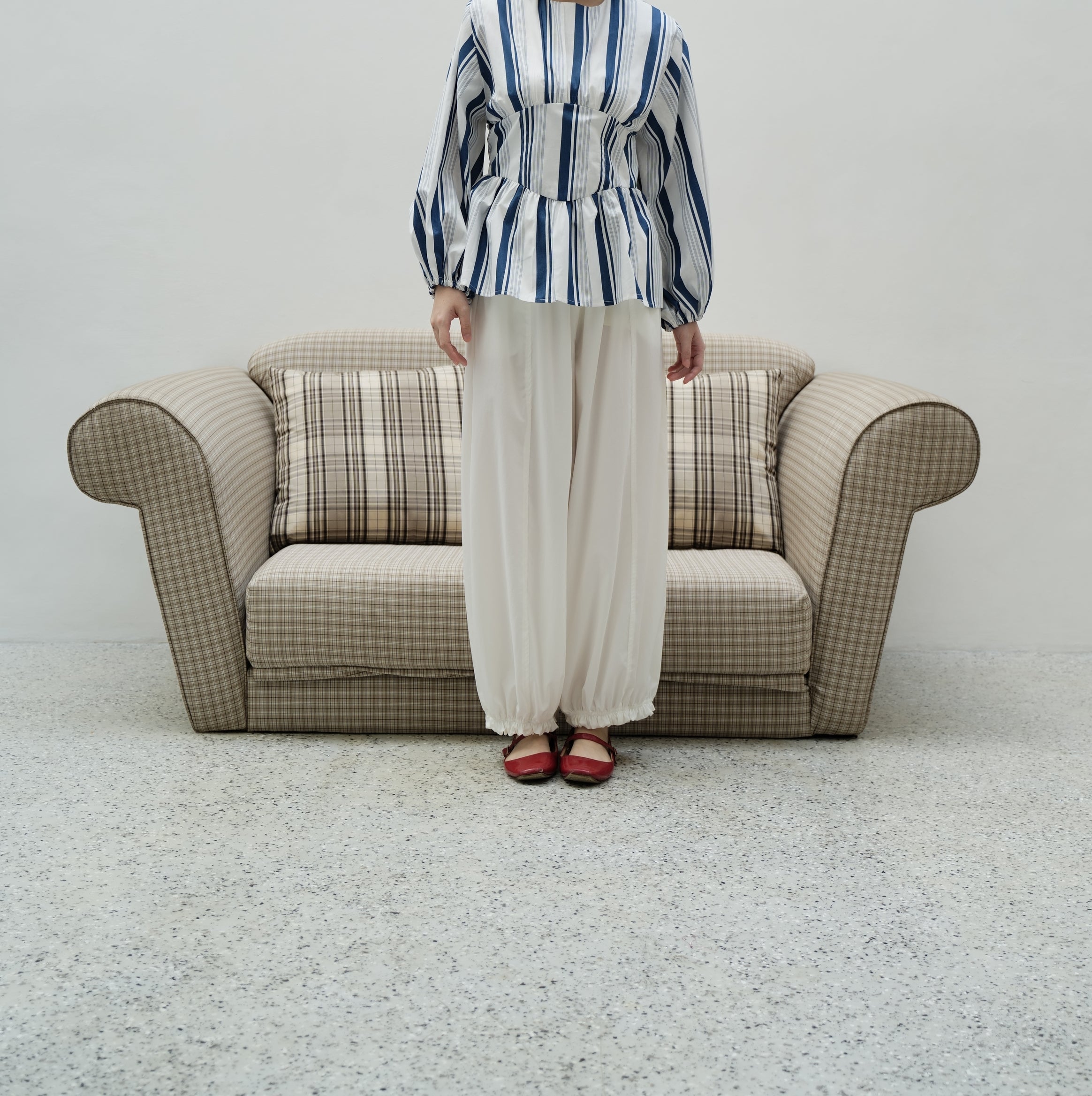 A woman stands in front of a beige armchair wearing a blue and white striped long-sleeve peplum top by BWBYAZ and white wide-leg pants, with red shoes, against a plain light gray wall.