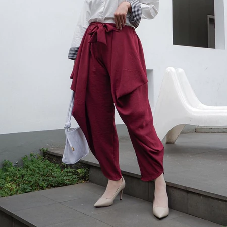 A woman standing in an outdoor setting wearing maroon rami pants by Abame and a white top.