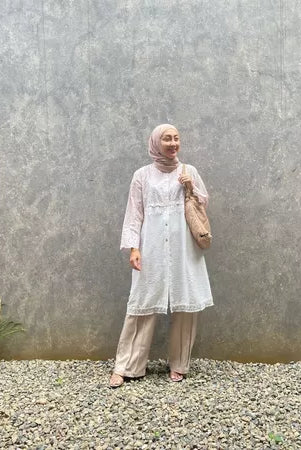 A woman standing outdoors wearing a cream-colored tunic with long sleeves, round neckline, and long pants. She has a bag and is posing against a gray wall with a textured background.
