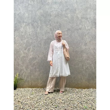 A woman standing outdoors wearing a cream-colored tunic with long sleeves, round neckline, and long pants. She has a bag and is posing against a gray wall with a textured background.
