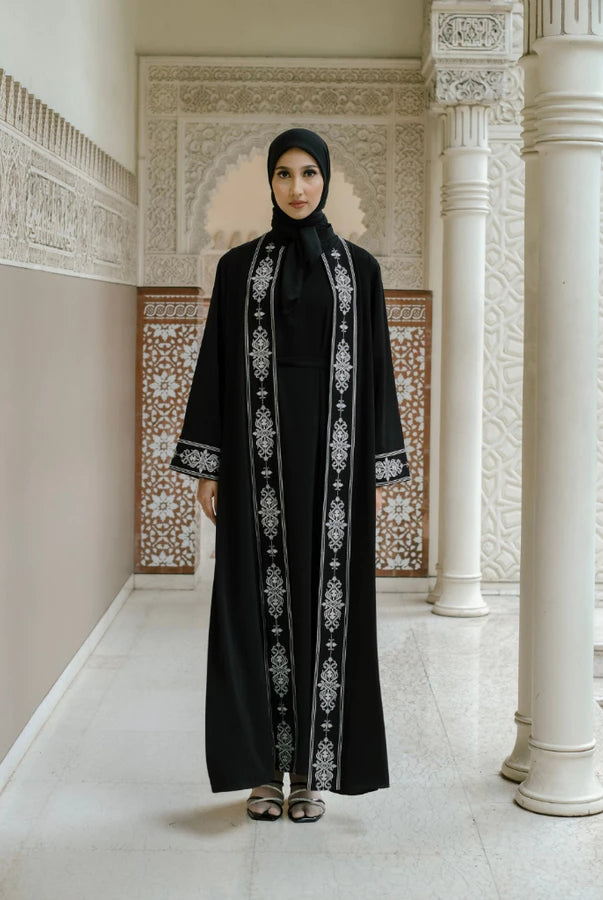 Woman in a black abaya with white embroidery by Tubita standing in an ornate hallway.