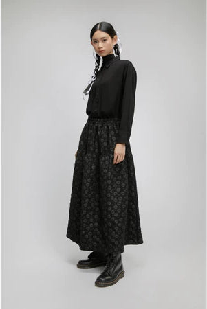 Person wearing a black turtleneck and patterned long black skirt by Yestoday on a gray background