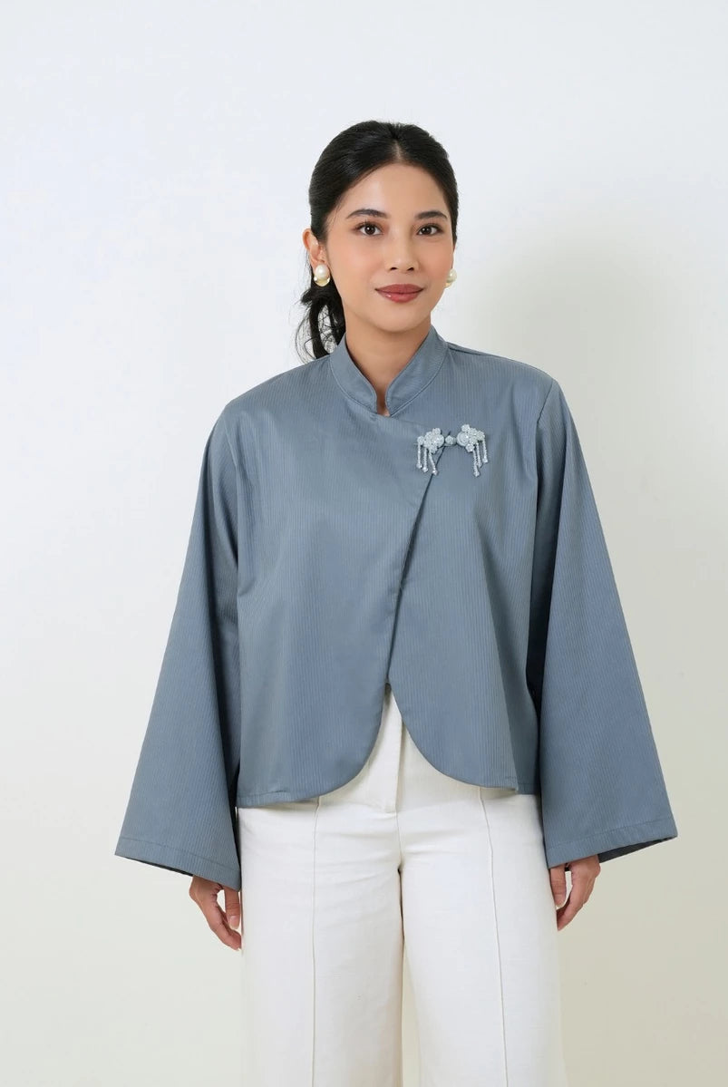 A woman wearing a slate blue cropped blouse by Studio Tui with long wide sleeves and a small white embroidered detail on the chest, paired with white pants, against a plain off-white background.