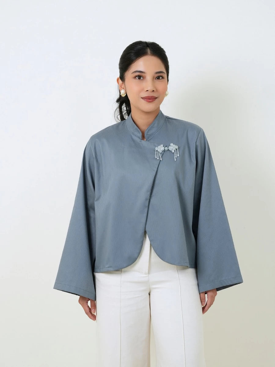A woman wearing a slate blue cropped blouse by Studio Tui with long wide sleeves and a small white embroidered detail on the chest, paired with white pants, against a plain off-white background.