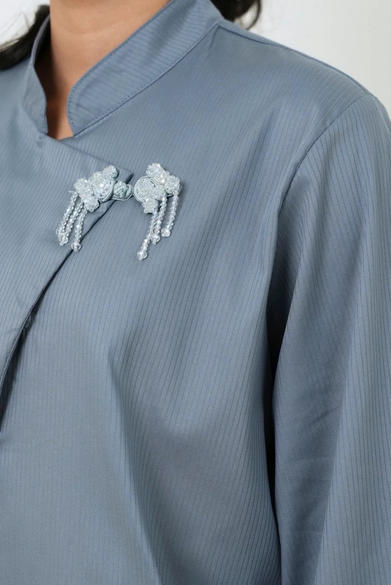 Close-up of a person wearing a light blue-gray long-sleeve blouse by Studio Tui with a mandarin collar and decorative silver beaded brooches on the chest.