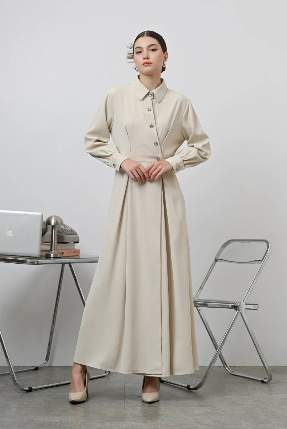A woman standing in an office setting wearing a beige long-sleeved Renata dress by Kasa Heritage with a fitted waist and silver buttons.