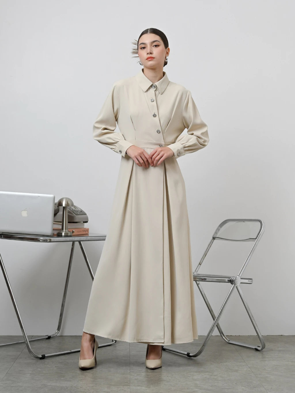 A woman standing in an office setting wearing a beige long-sleeved Renata dress by Kasa Heritage with a fitted waist and silver buttons.