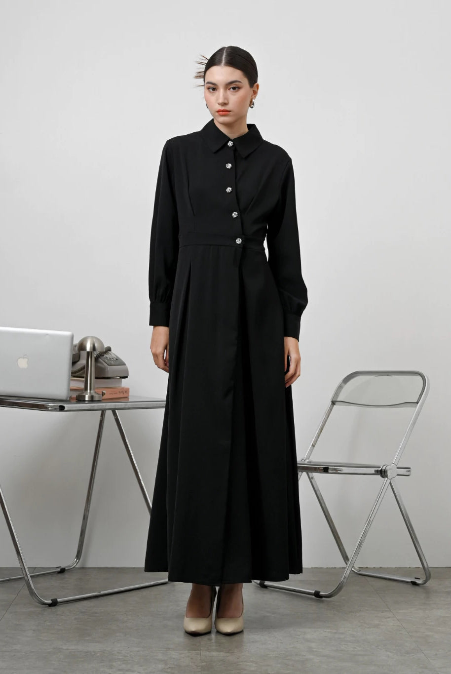 A woman standing in an office setting wearing a black long-sleeved Renata dress by Kasa Heritage with a fitted waist and silver buttons.