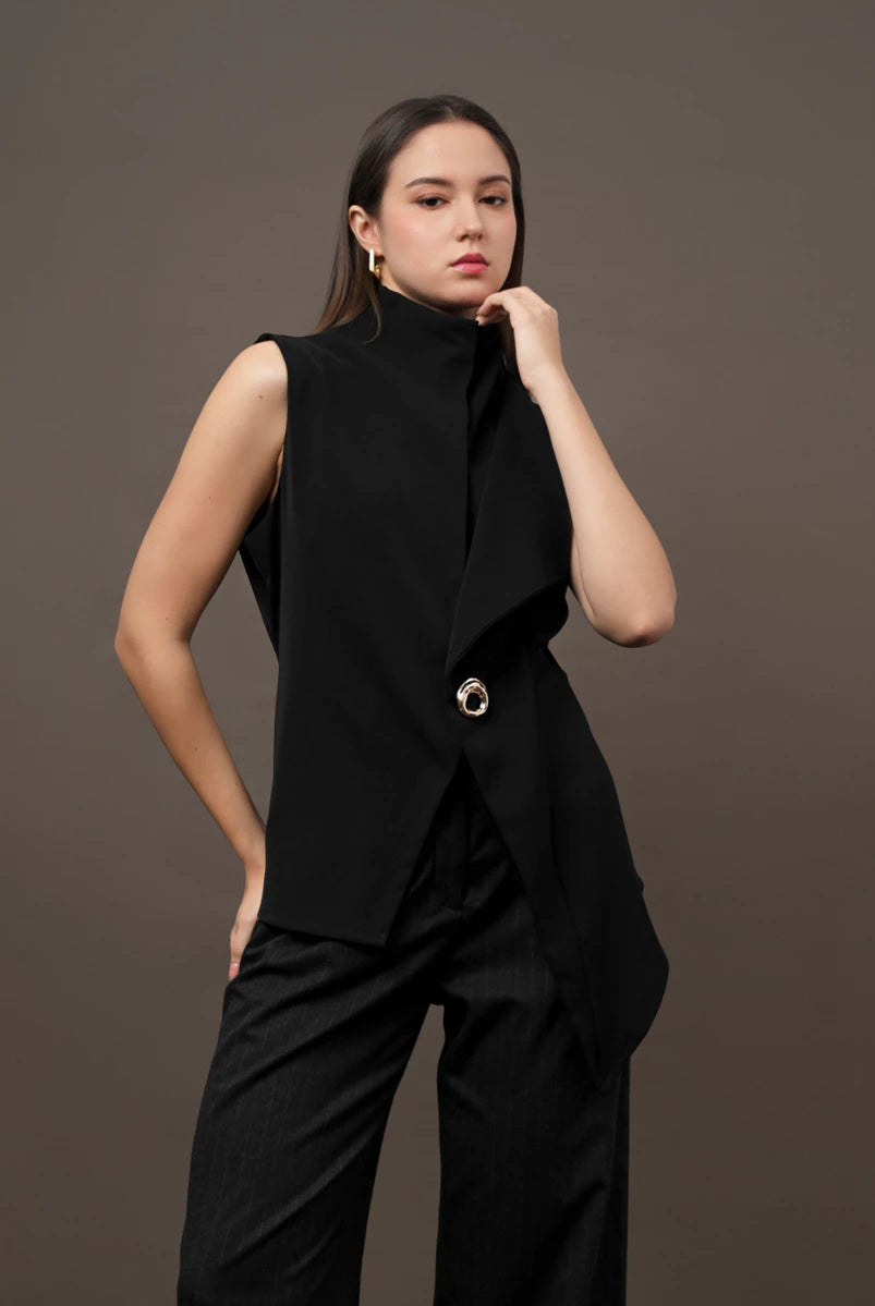 A woman modeling a black sleeveless wrap-style vest by Kasa Heritage with a high mock neck and a single prominent gold-tone button, paired with black wide-leg trousers, against a solid dark brown studio background.