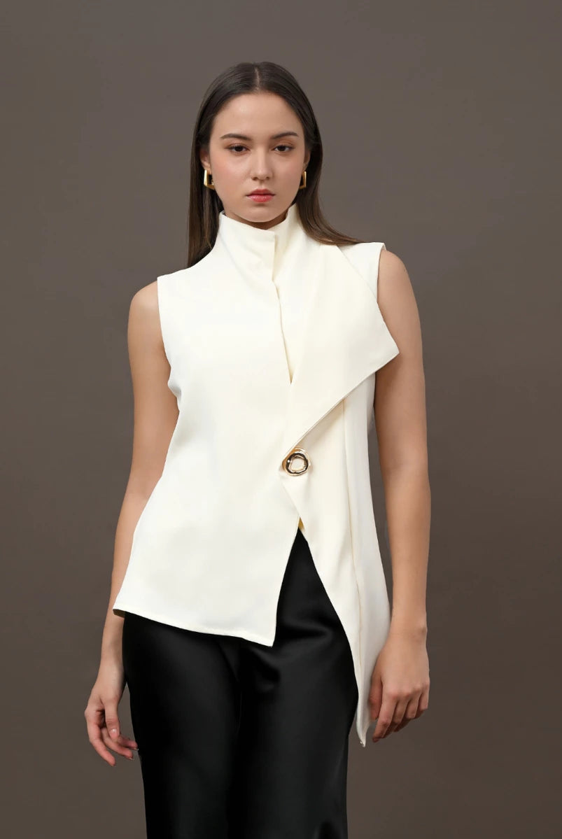 A model wearing a sleeveless high-neck wrap vest in off-white by Kasa Heritage with a gold-tone circular accent, paired with black pants, against a solid dark gray studio background.