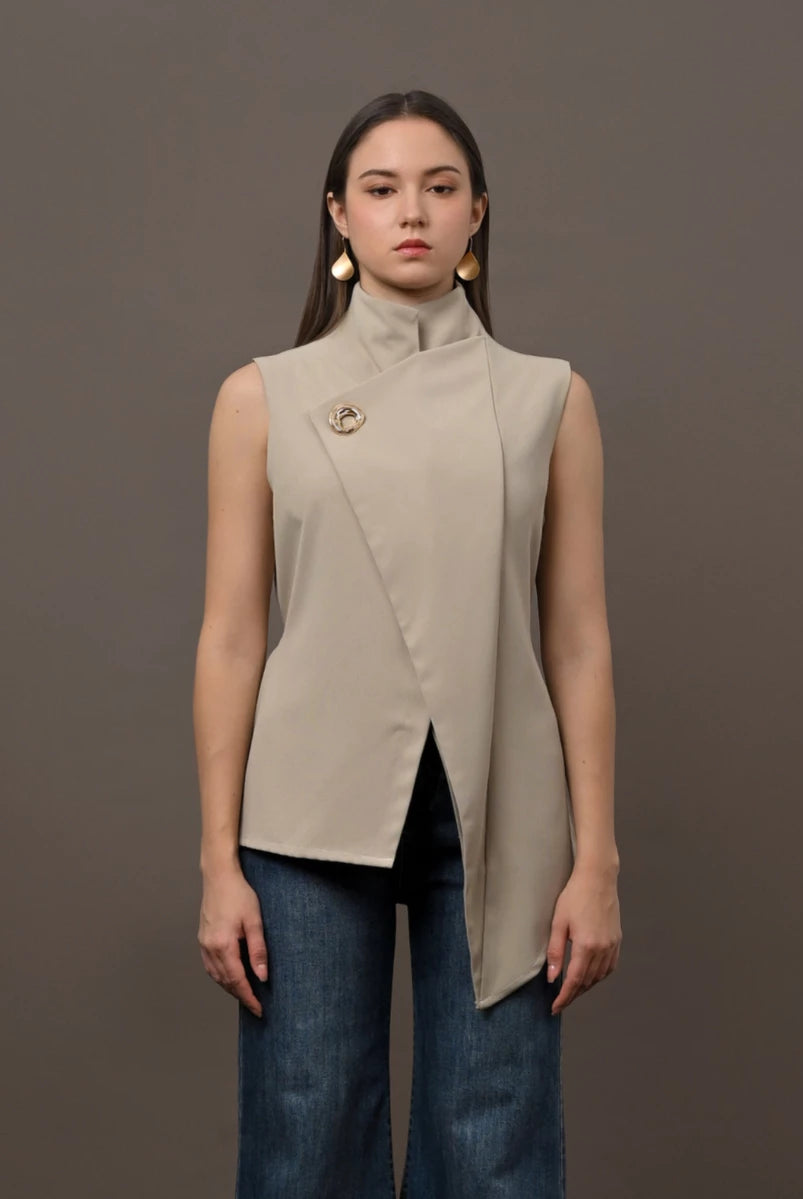 A woman modeling a sleeveless beige wrap-front vest by Kasa Heritage with a high mock neck and a decorative gold pin on the chest, paired with dark blue jeans, against a solid dark gray studio background.