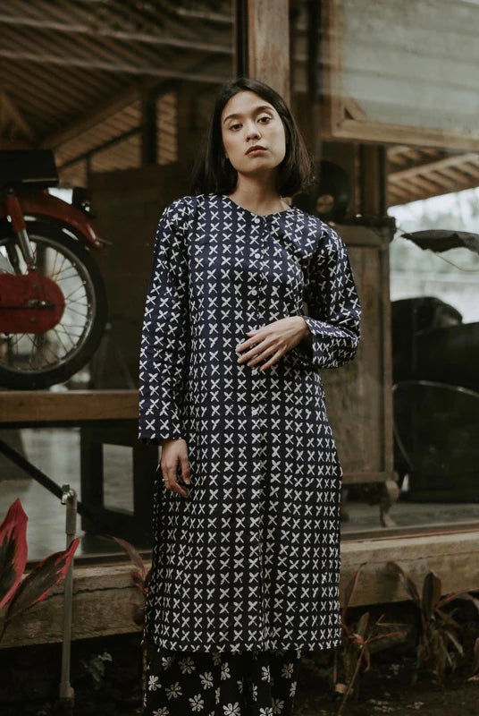 A woman wearing a long-sleeved navy blue tunic with a white geometric pattern, standing in a rustic wooden structure with a red motorcycle in the background.