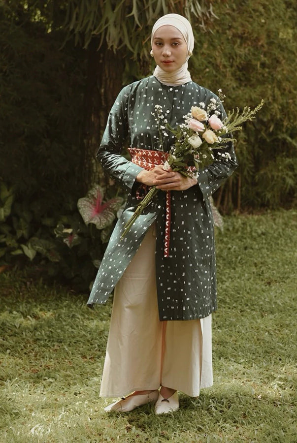 A woman wearing a long-sleeve tunic in a dark green patterned fabric with a red patterned sash, paired with a cream-colored skirt and a white hijab, standing in a grassy garden while holding a bouquet of pink and white flowers.