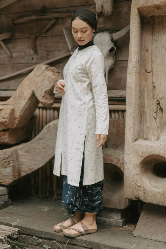 A woman wearing a long white patterned tunic with dark patterned pants and flat sandals, standing in front of a rustic wooden wall with carved wood and a bull skull.