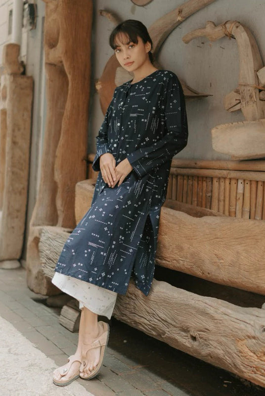 A woman wearing a navy blue long-sleeve tunic with a white celestial print, leaning against a rustic wooden bench in an outdoor setting.