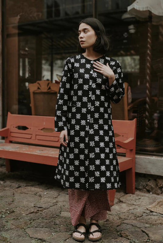 A woman wearing a black long-sleeve tunic dress with a white floral pattern, paired with a red and white checkered skirt, standing in front of a rustic red wooden bench and shop window.