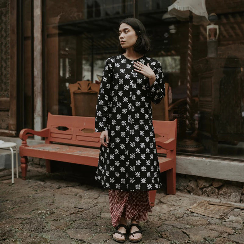 A woman wearing a black long-sleeve tunic dress with a white floral pattern, paired with a red and white checkered skirt, standing in front of a rustic red wooden bench and shop window.