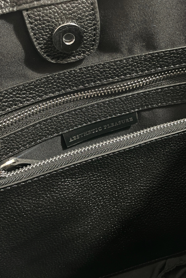 Close-up of a black grain leather bag with a zipper, featuring the brand name 'Aesthetic Pleasure'.