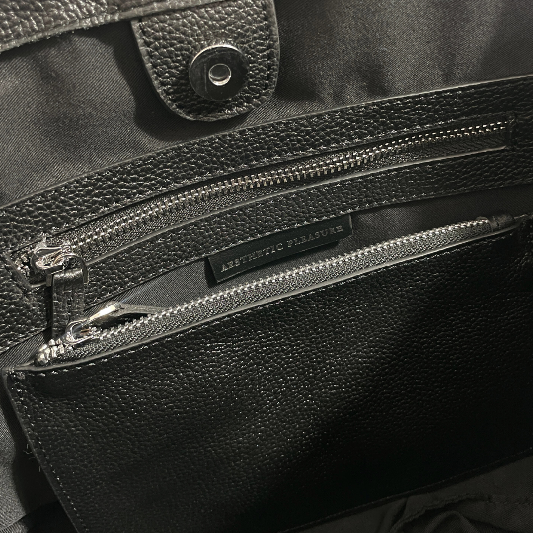 Close-up of a black grain leather bag with a zipper, featuring the brand name 'Aesthetic Pleasure'.