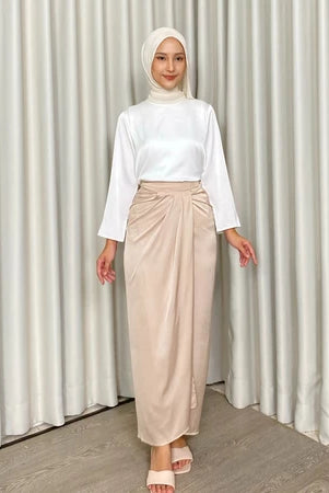 Woman wearing a white top and beige skirt by Kartinis Label against a white curtain background